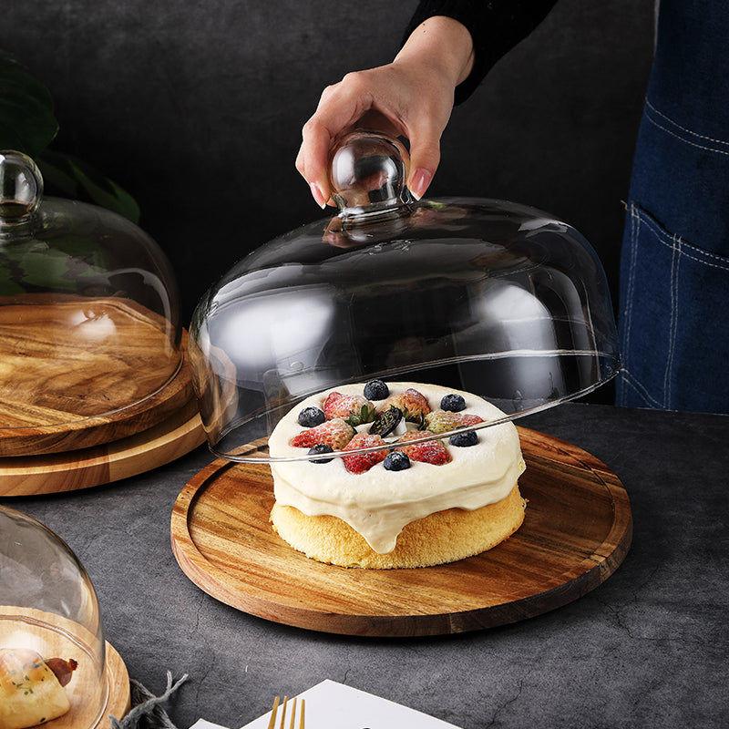 Hand lifting the glass dome off a cake on a wood cake plate