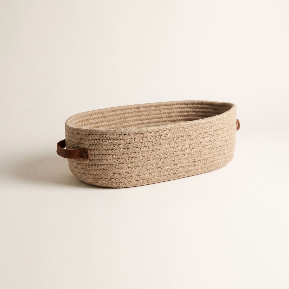Beige oval woven rope basket with brown handle tabs on a cream backdrop