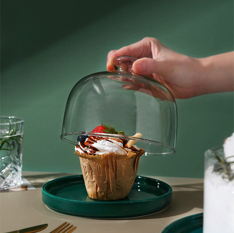 Hand lifting a glass dome off a green plate with a cupcake