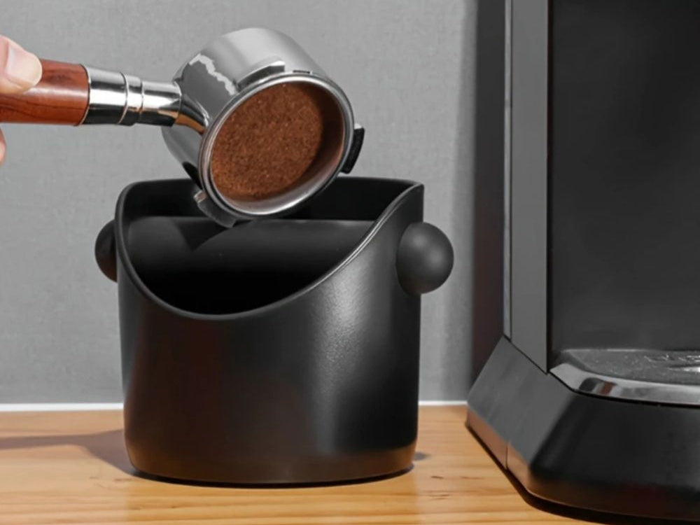 Knocking a used espresso puck into the black knock box beside a machine