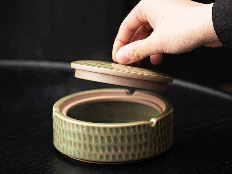 Hand lifting the lid off a green ceramic ashtray on a dark wood table