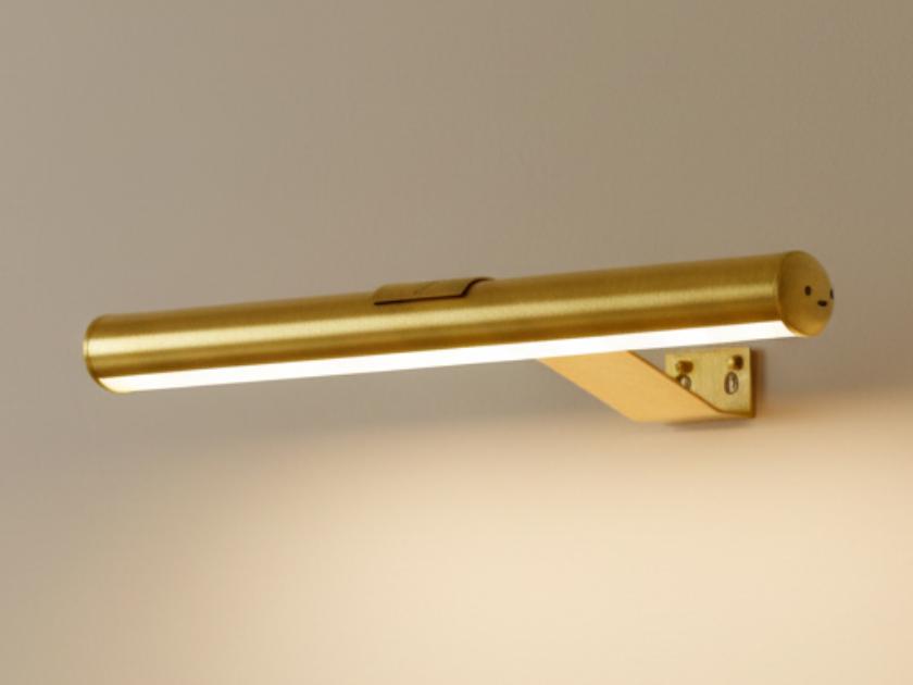 Gold picture light bar on its angled wall bracket, switched on with a warm glow