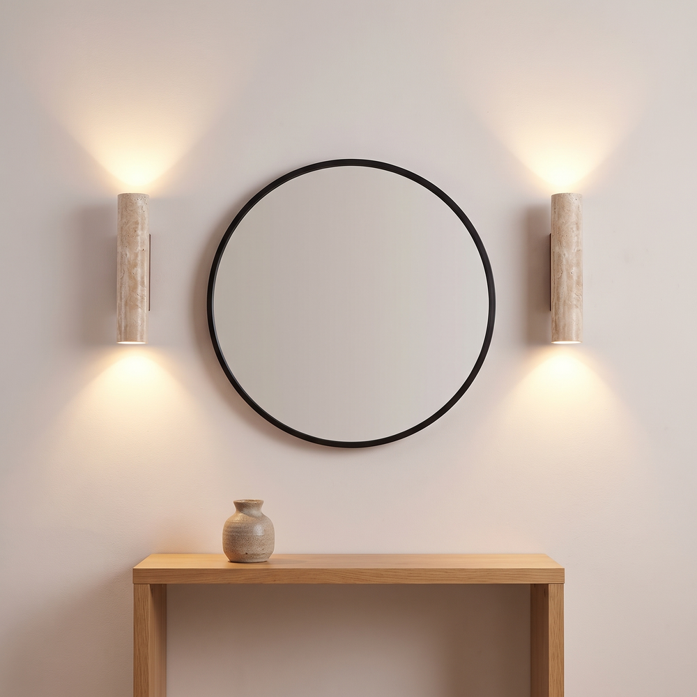 Two travertine wall sconces flanking a round mirror above an oak console, each washing the wall with warm light above and below